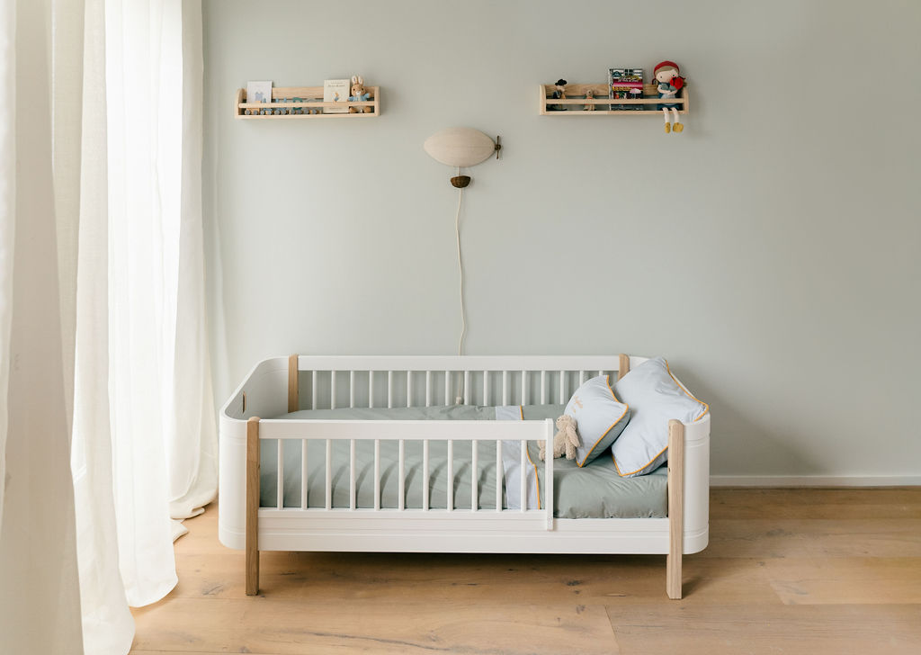 A softly lit children's room with a white bed dressed in sage and pale blue linens with yellow piping